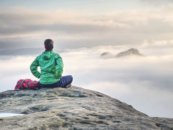 Rear view of hiker woman on top of mountain peak sit in silence. thick fog covered world