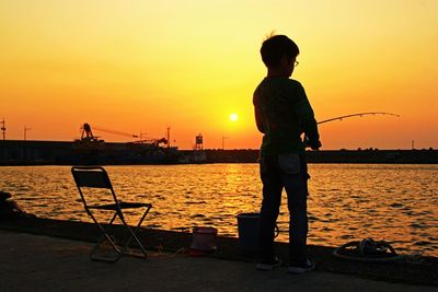 Rear view of boy fishing in lake against orange sky