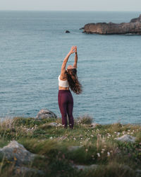 Back view of young fit female with long wavy hair in sportswear performing mountain with arms up and backbend asana while practicing yoga on grassy hill at seaside