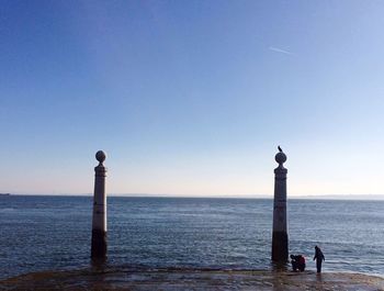 People by tall bollards on shore against blue sky