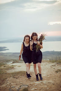 Two woman in black dresses walk on the mountain with a bouquet of flowers in summer at sunset