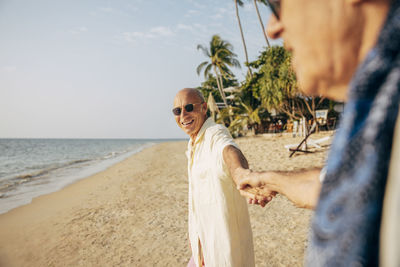 Happy senior man holding hands of boyfriend while standing on beach at sunny day