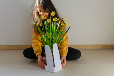 Child with long dark hair and yellow top adorning daffodil plant pot with rabbit ears for easter