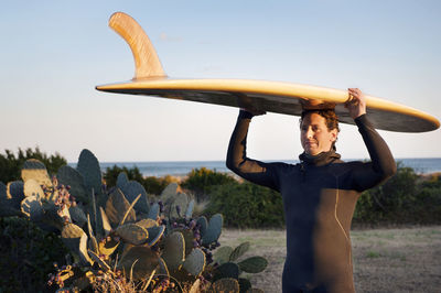 Surfer carrying surfboard on head while standing on grassy field against clear sky