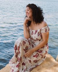 Beautiful young woman sitting on rock against sea