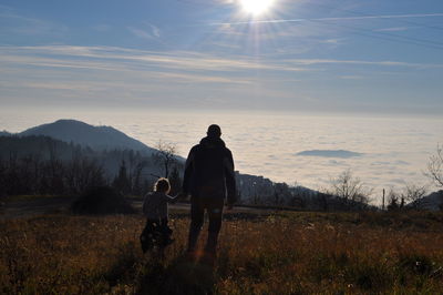 Rear view of father and daughter walking on grassy field by cloudscape against sky