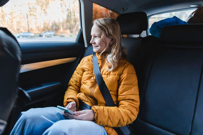 Content teen girl sitting in car on passenger seat and browsing mobile phone while looking out of window