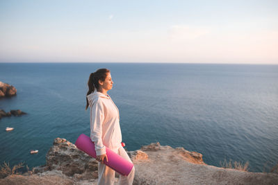 Smiling woman hold yoga mat wear sportswear hoodie and pants stand on top of rock over sea