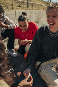 Woman painting fingernail of happy man sitting by friends in park on sunny day