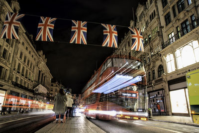 Women by blurred motion of bus with british flags hanging in city at night