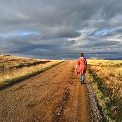 Rear view of young woman walking along dirt road in green field against cloudy sky