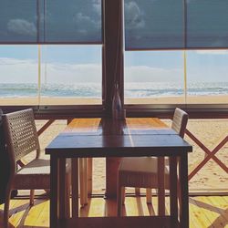 Empty chairs and table by sea against sky seen through window