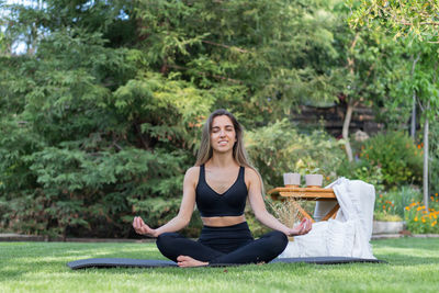 Front view of young woman practicing lotus pose and chilling on garden while enjoying doing yoga