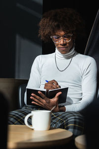 Confident young african american male student with curly hair in stylish outfit and eyeglasses taking notes in copybook while preparing for exam in modern cafe on sunny day