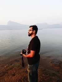 Side view of young man with camera standing at lakeshore against clear sky