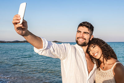 Portrait of smiling young man using mobile phone against sea