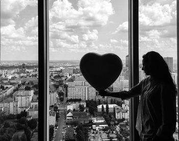 Woman holding heart decoration while looking at cityscape through window