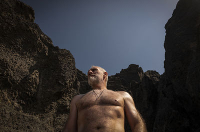 Low angle view of portrait of shirtless adult man on beach against rock