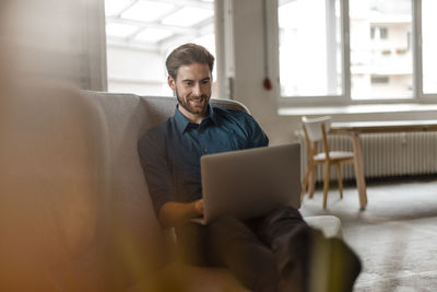 Portrait of young freelancer sitting on couch in a loft using laptop