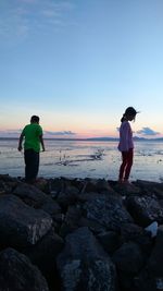 Siblings standing on rock at beach against sky during sunset