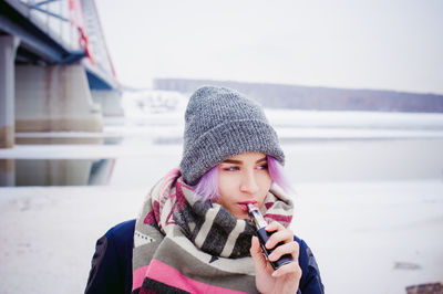 Young woman smoking while standing by bridge against sky during winter