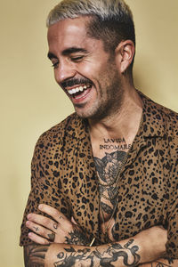 Portrait of cheerful fashionable male model with tattoos wearing trendy leopard shirt standing against beige background with closed eyes