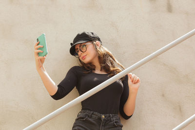 Young woman taking selfie standing against wall