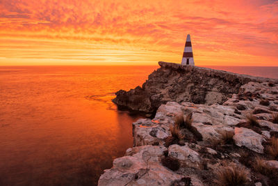 Lighthouse on rock by sea against sky during sunset