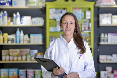 Portrait of female pharmacist with clipboard standing against shelf in store
