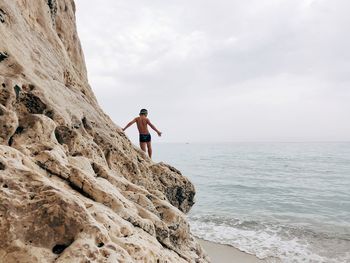 Rear view of shirtless boy standing on rock at beach against sky