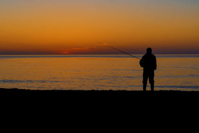 Silhouette man fishing in sea against orange sky