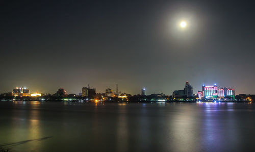 Illuminated buildings by river against sky at night