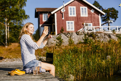 Smiling woman doing video conference on mobile phone while sitting over cushion on pier against house
