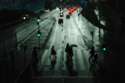 High angle view of silhouette people crossing road seen through wet window at night