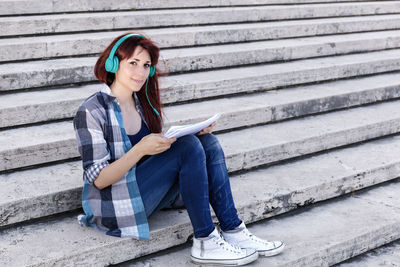 Portrait of woman reading papers while listening to music through headphones at steps