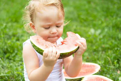 Funny smiling kid boy in white bodysuit eating watermelon at green lawn