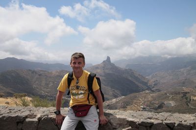 Portrait of smiling hiker sitting on retaining wall by mountains against sky