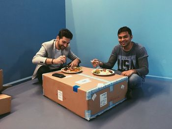 Happy men eating meal on cardboard box against wall at home