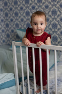 Baby girl in red overall standing at the railing of her bed and looking at camera. vertical format