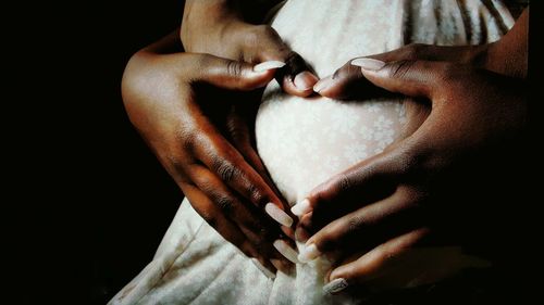 Close-up of couple hands making heart shape on pregnant abdomen against black background