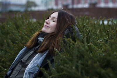 Smiling woman with closed eyes leaning on plants at park during winter