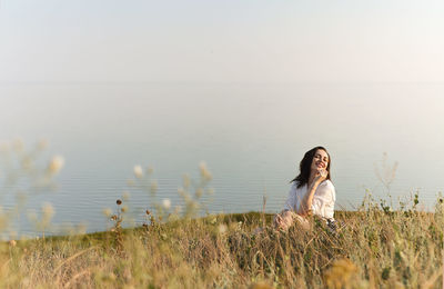 Young woman on grass at shore against clear sky