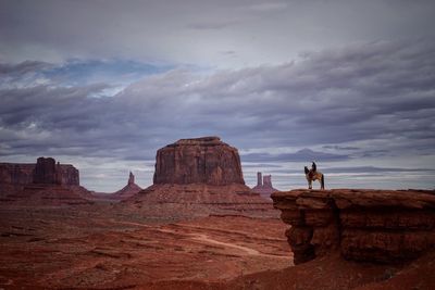 Man with horse on cliff by rock formations against cloudy sky at monument valley