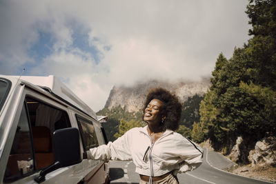 Smiling woman with eyes closed standing next to van during road trip