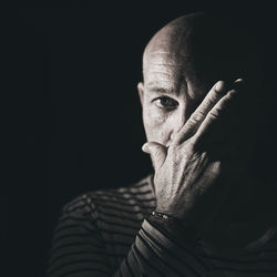 Close-up portrait of bald man covering mouth against black background