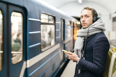 Man holding his smartphone in hands, listen music with headphones and wait the train.