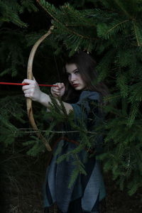 Young woman holding aiming bow while standing in forest