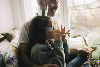 Girl wearing crown and glasses enjoying with ribbon while sitting on father at home