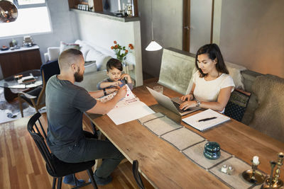 Mother working on laptop while father assisting son in doing homework at dining table