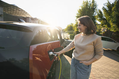 Happy woman charging electric car at roadside on sunny day